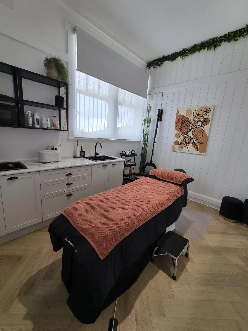 A massage room with a massage table draped in a coral-colored blanket, a step stool at the foot, and a white wall with artwork and shelves containing supplies.