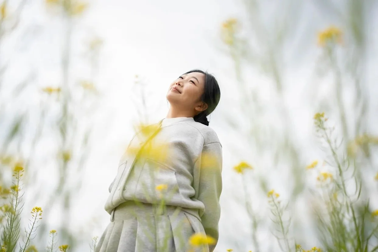 A woman standing in soft spring sunlight with eyes closed, embodying calm, grounded presence.