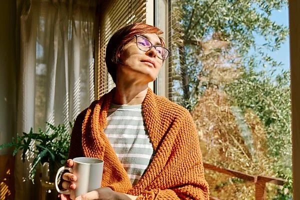 Woman sitting quietly by a window in soft morning light holding a mug, reflecting in a calm and contemplative moment.
