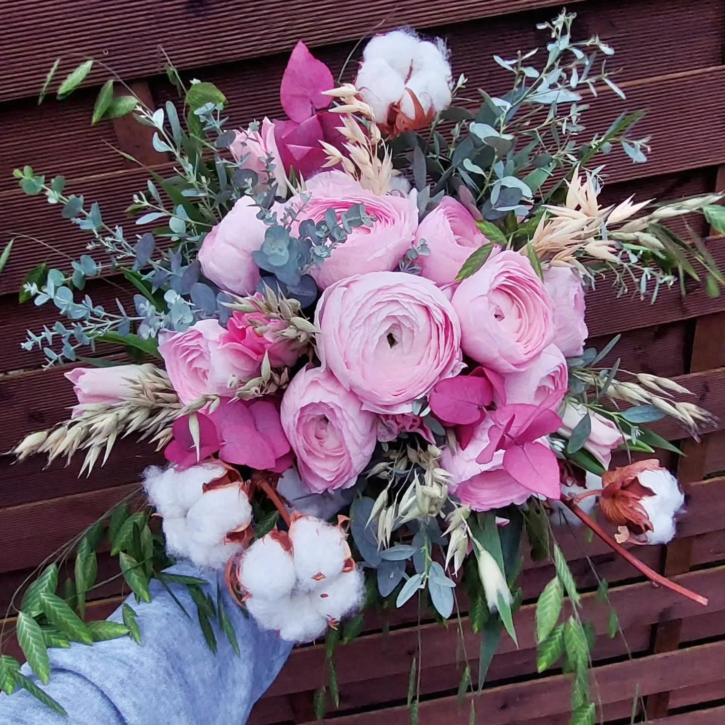 Un bouquet de fleurs avec des roses roses, des boules de coton, des épis de blé et des feuillages verts, tenu par une personne portant un vêtement gris, avec un fond en bois.