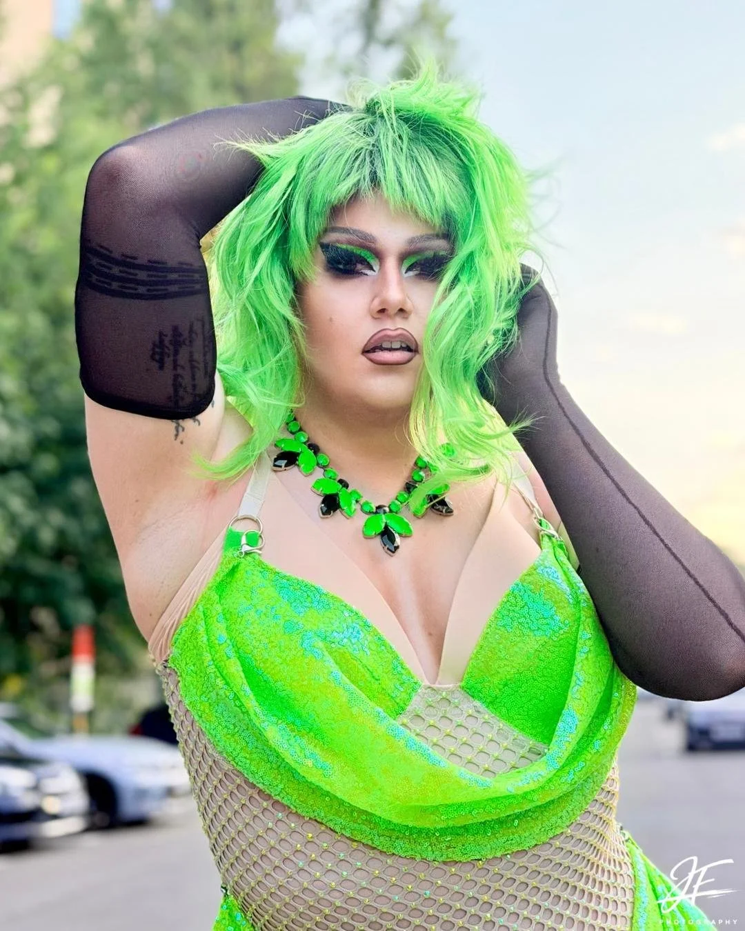 Person with bright green hair in a messy style, wearing a neon green and beige dress with black fishnet gloves, a black and green necklace, and makeup with bold eye shadow, standing outdoors with trees and parked cars in the background.