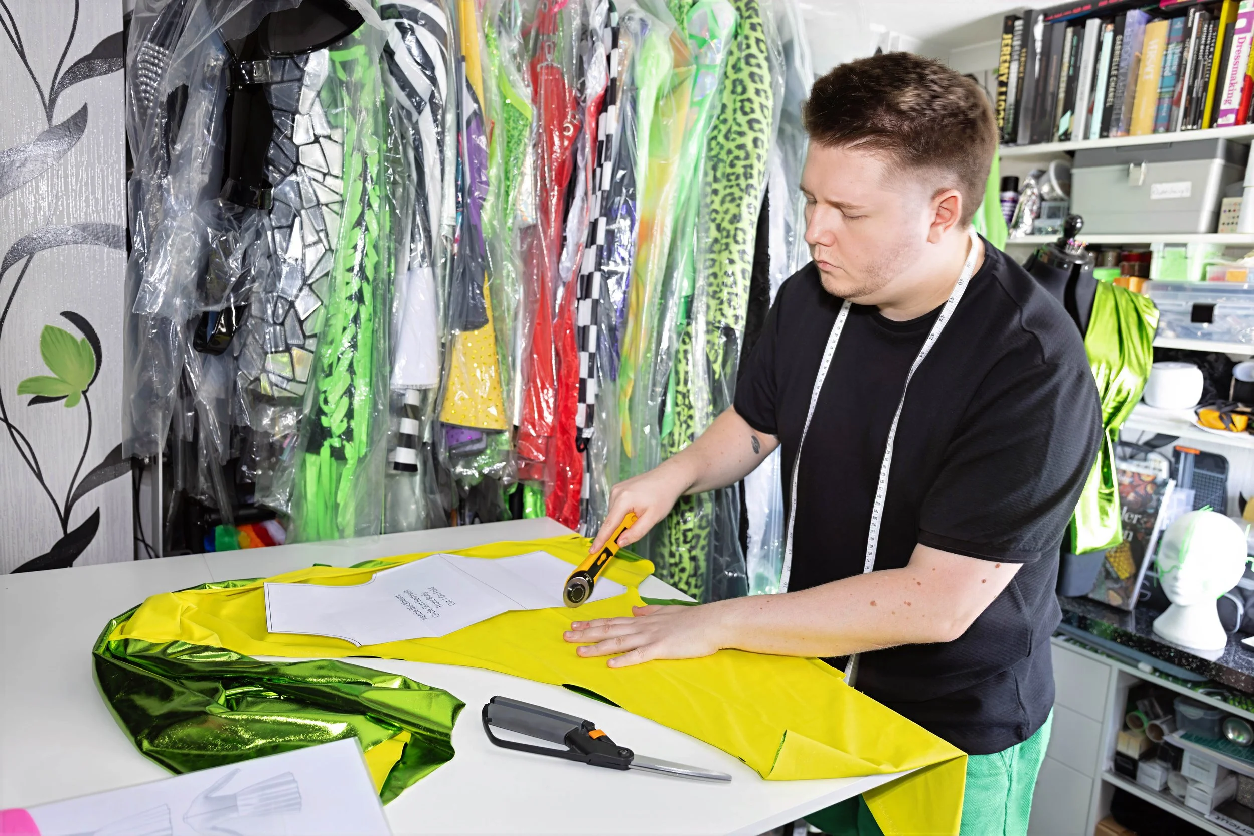 A person in a workshop or craft room using a hot glue gun on a yellow, metallic, possibly inflatable item. The workspace is organized with shelves holding books, tools, and supplies. Colorful fabrics or materials are hanging on a wall in the background.