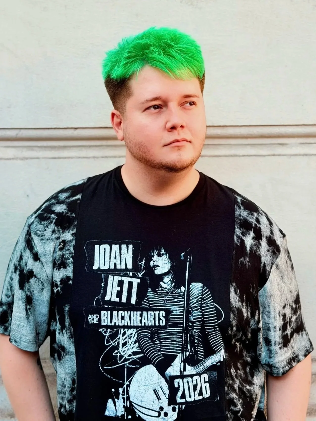 Young man with short brown hair and green dyed hair standing against a beige wall, wearing a black Joan Jett concert T-shirt and a tie-dye patterned jacket.