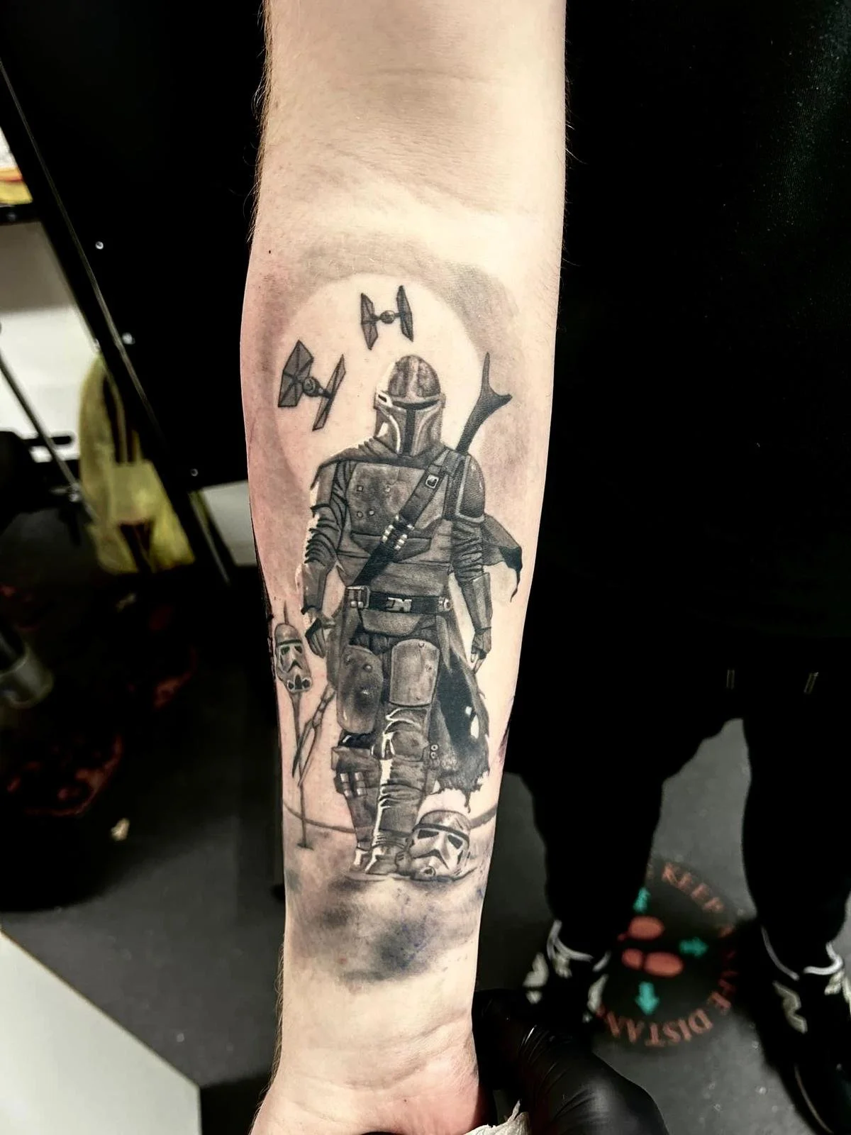 Black and gray tattoo of The Mandalorian character walking with four Star Wars TIE fighters flying above, a skull mask on the ground, and a TIE fighter helmet next to the character.
