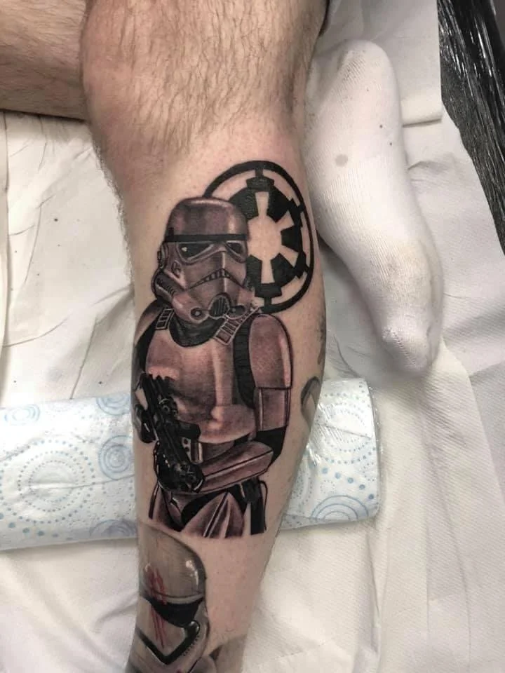 Tattoo of a Stormtrooper holding a gun on a person's calf, with the Galactic Empire emblem in the background.