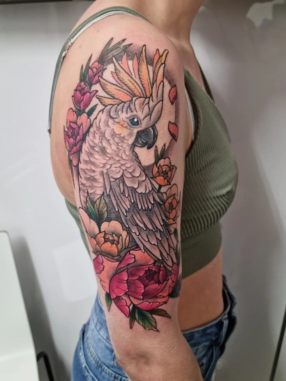 A colorful tattoo of a cockatoo bird with pink, orange, and gray feathers, surrounded by pink and orange flowers, on a woman's upper arm and shoulder.