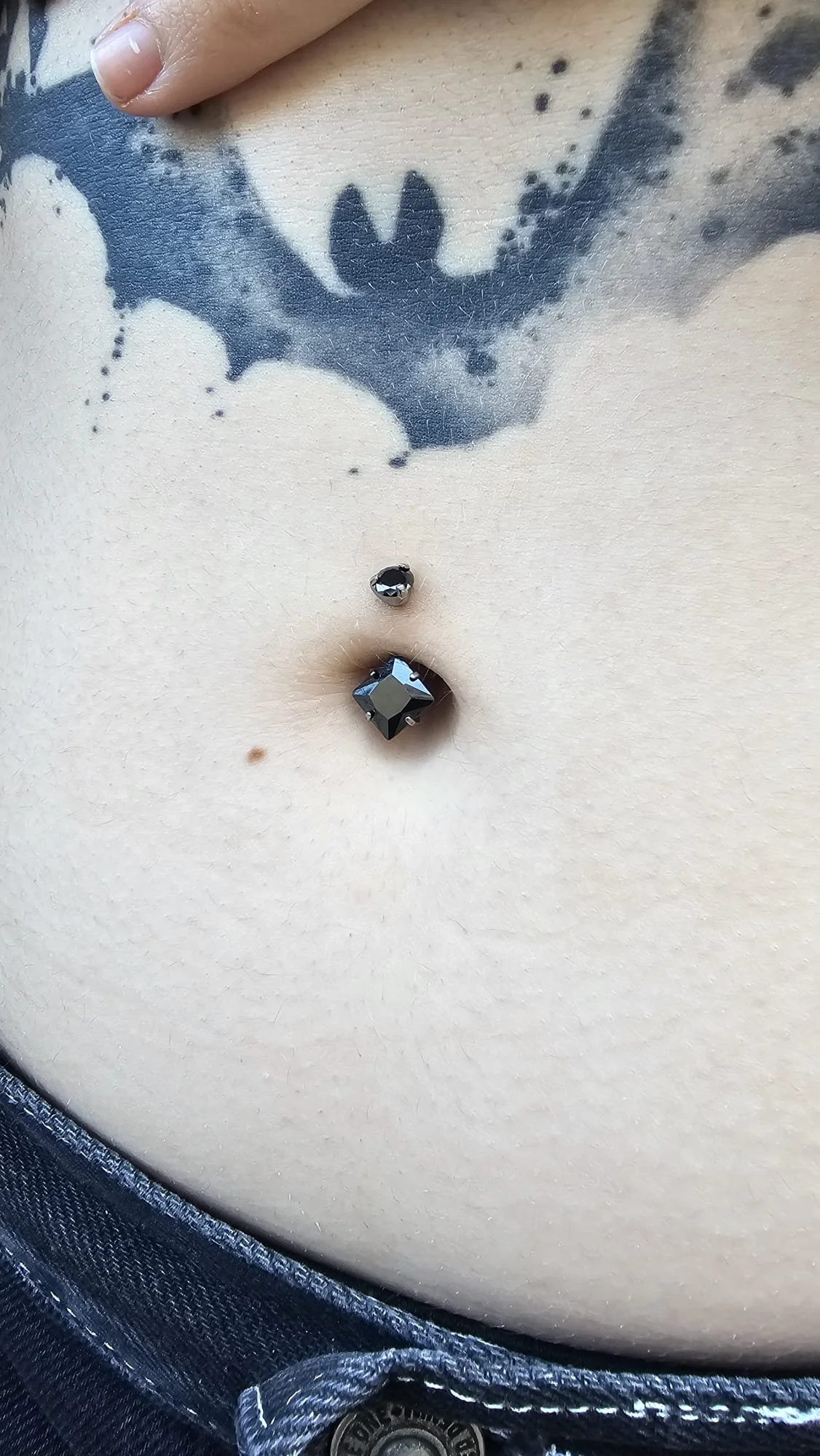 Close-up of a person's navel with a black jewelry piercing and part of a tattooed chest showing a bat wing.