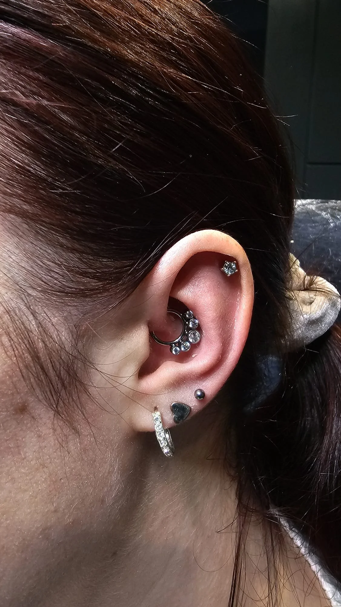 Close-up of a person's ear with multiple earrings, including a small hoop with rhinestones, a black heart-shaped earring, a small black ball, and a silver hoop with crystals.
