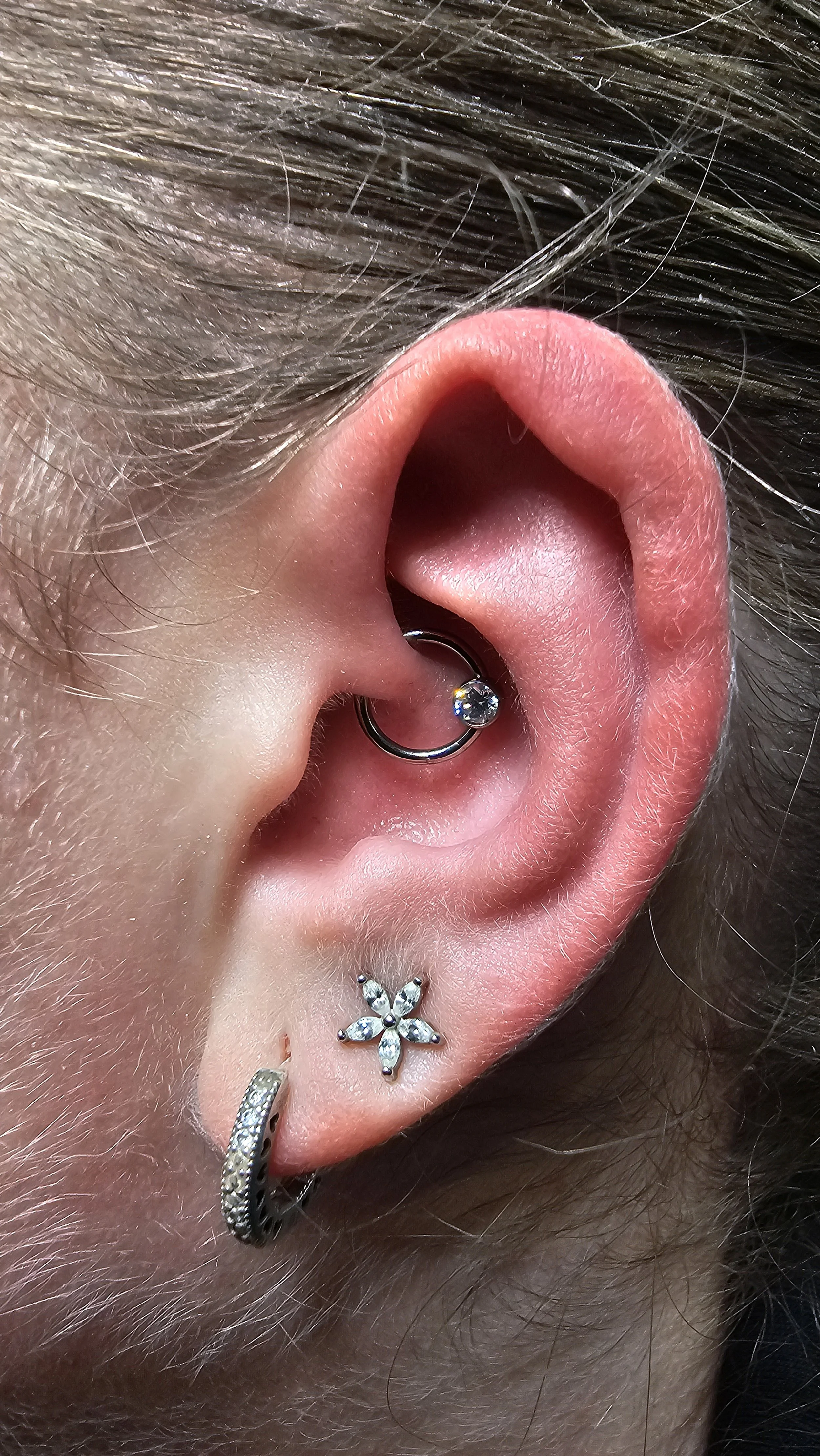 Close-up of a human ear with multiple piercings, including a silver hoop with a gemstone, a flower-shaped earring with clear stones, and a small hoop with silver details.