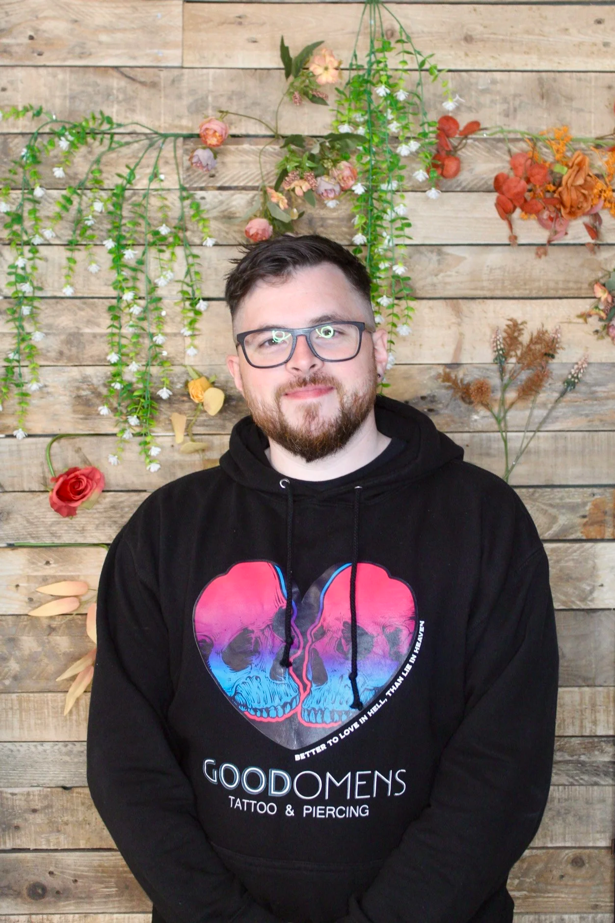 A man with glasses and a beard wearing a black hoodie with a colorful skull graphic and text, standing in front of a wooden wall decorated with hanging flowers.