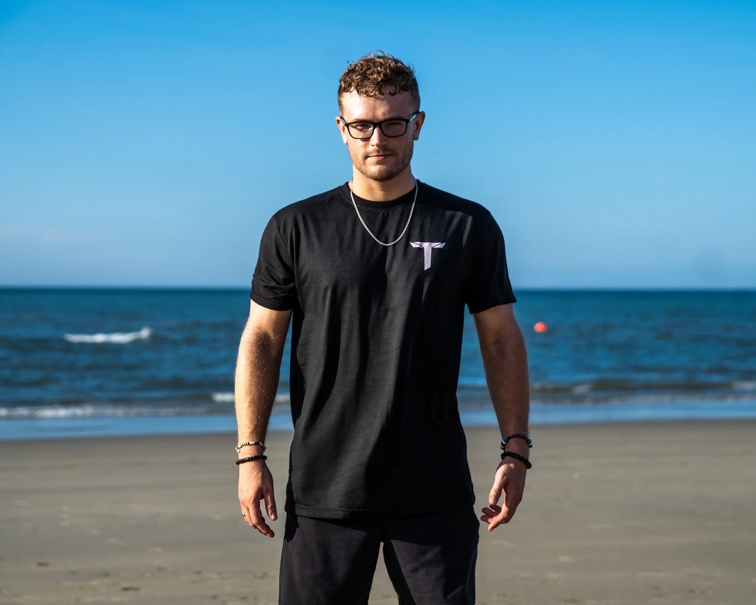A man standing on a beach with the ocean and clear blue sky in the background. He is wearing a black T-shirt with a Tesla logo, bracelets, and glasses.
