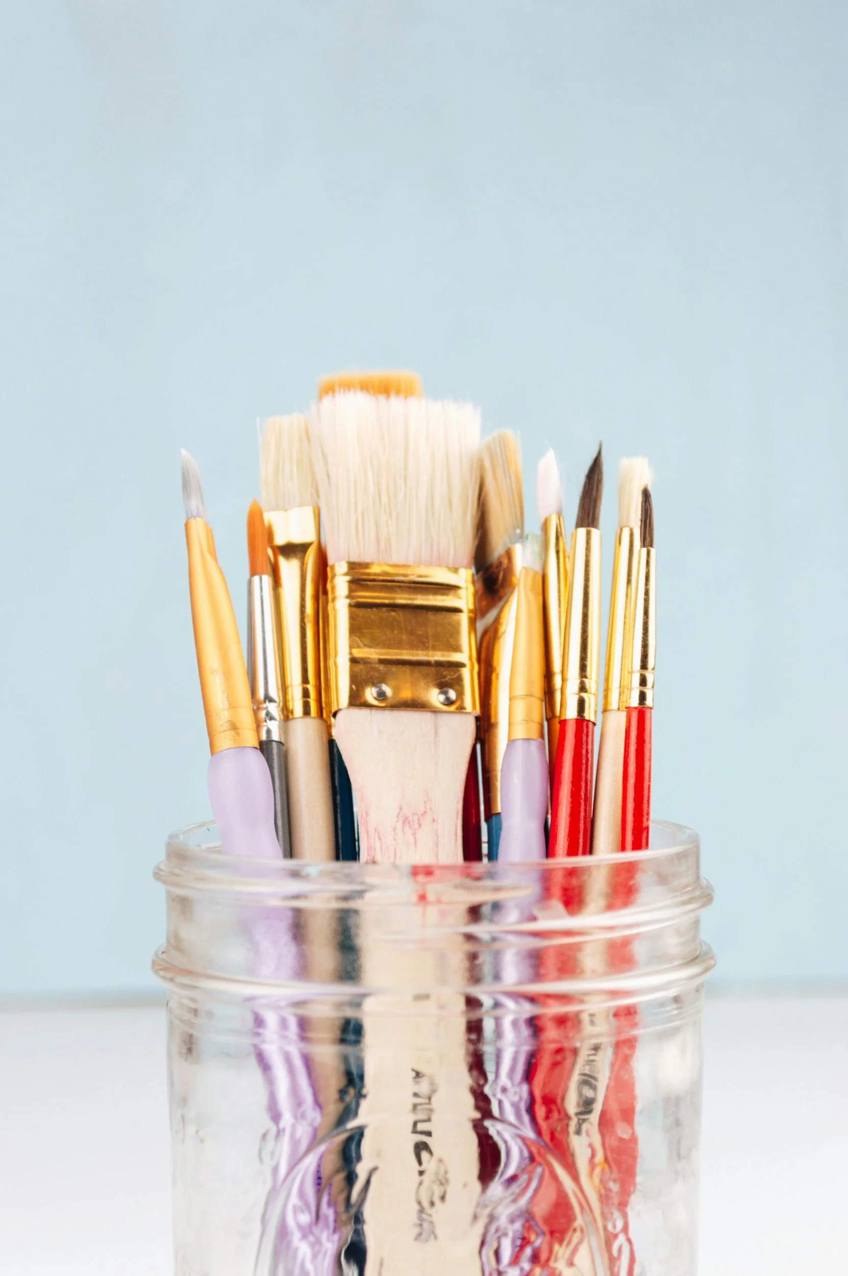 A glass jar filled with various paintbrushes with white, purple, and red handles, some with gold ferrules, against a light blue background.