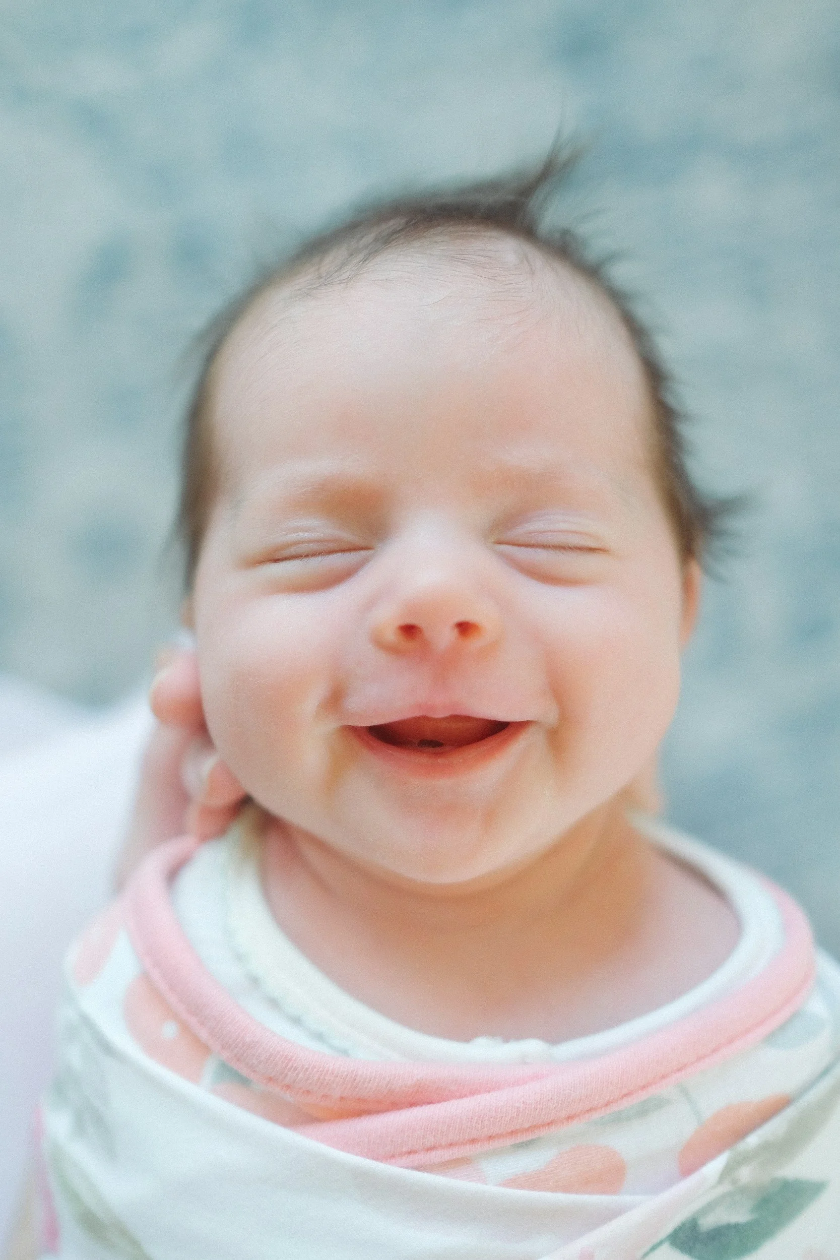 A smiling baby with closed eyes and a happy expression, wearing a light-colored outfit with pink accents.