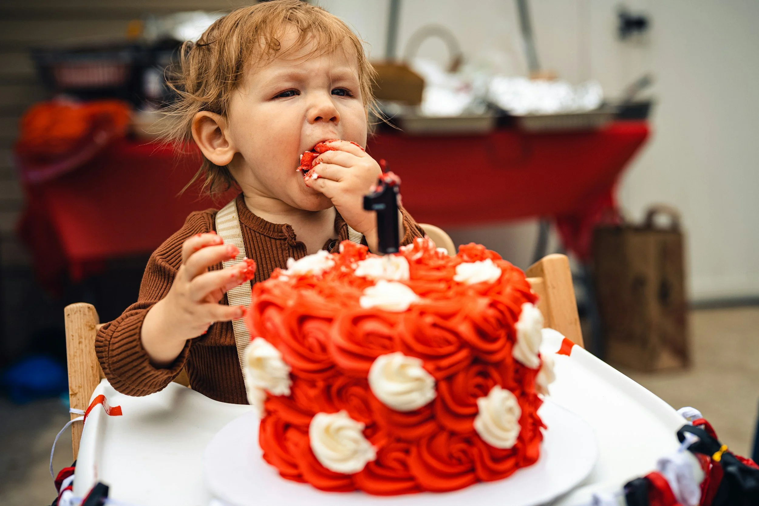 A young child with curly hair eating a slice of orange-colored cake decorated with white swirls, sitting at a table with a large birthday cake in front of them.