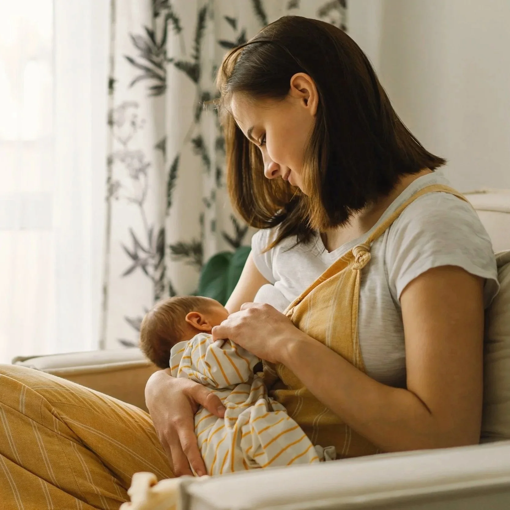 A mother with shoulder-length brown hair breastfeeds a newborn baby while sitting on a sofa in a cozy, well-lit room with patterned curtains in the background.