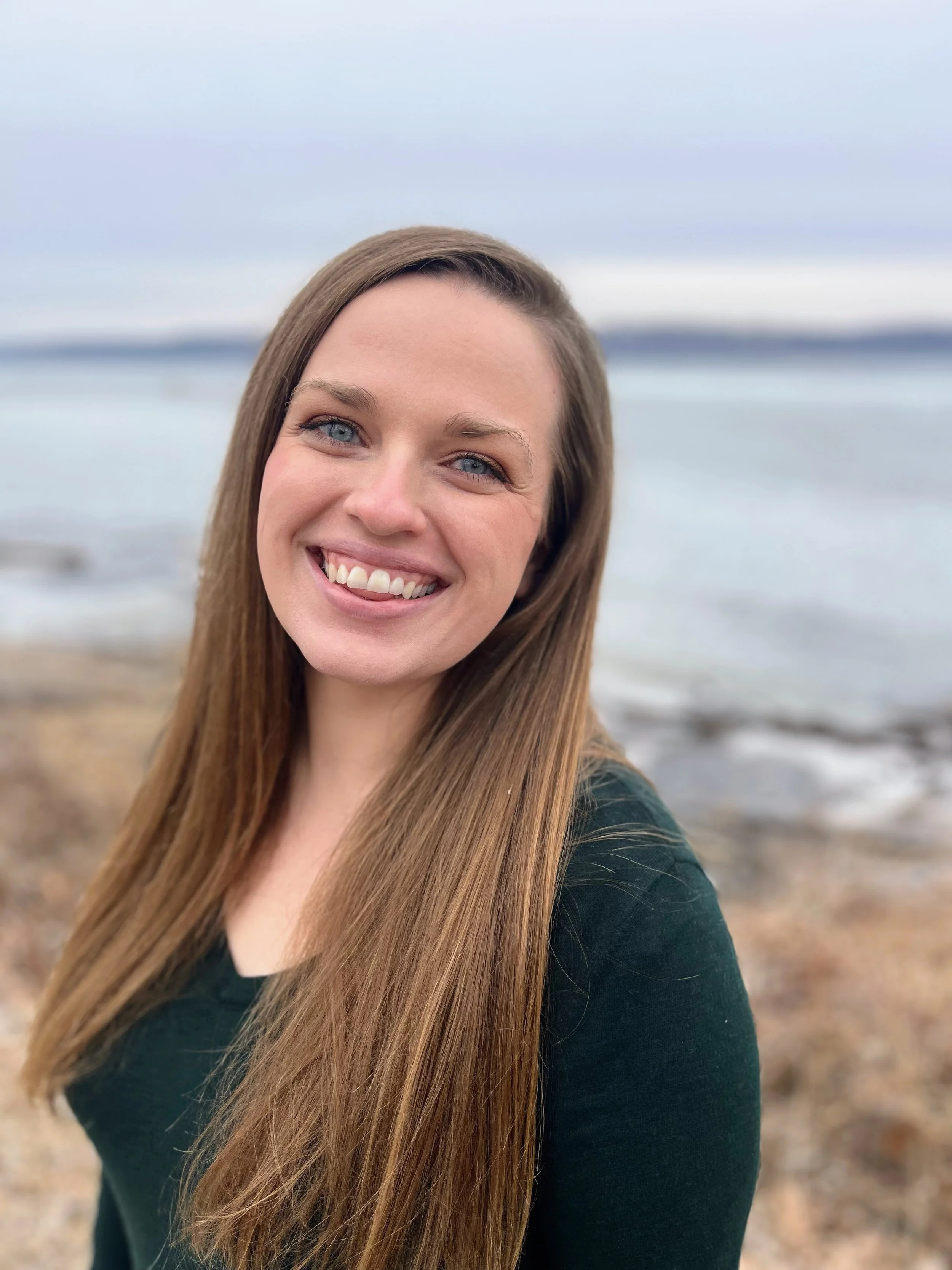 Birth and Postpartum Doula, Stephanie Cook, with straight brown hair and blue eyes smiling outdoors near a body of water, with a rocky beach and a cloudy sky in the background.