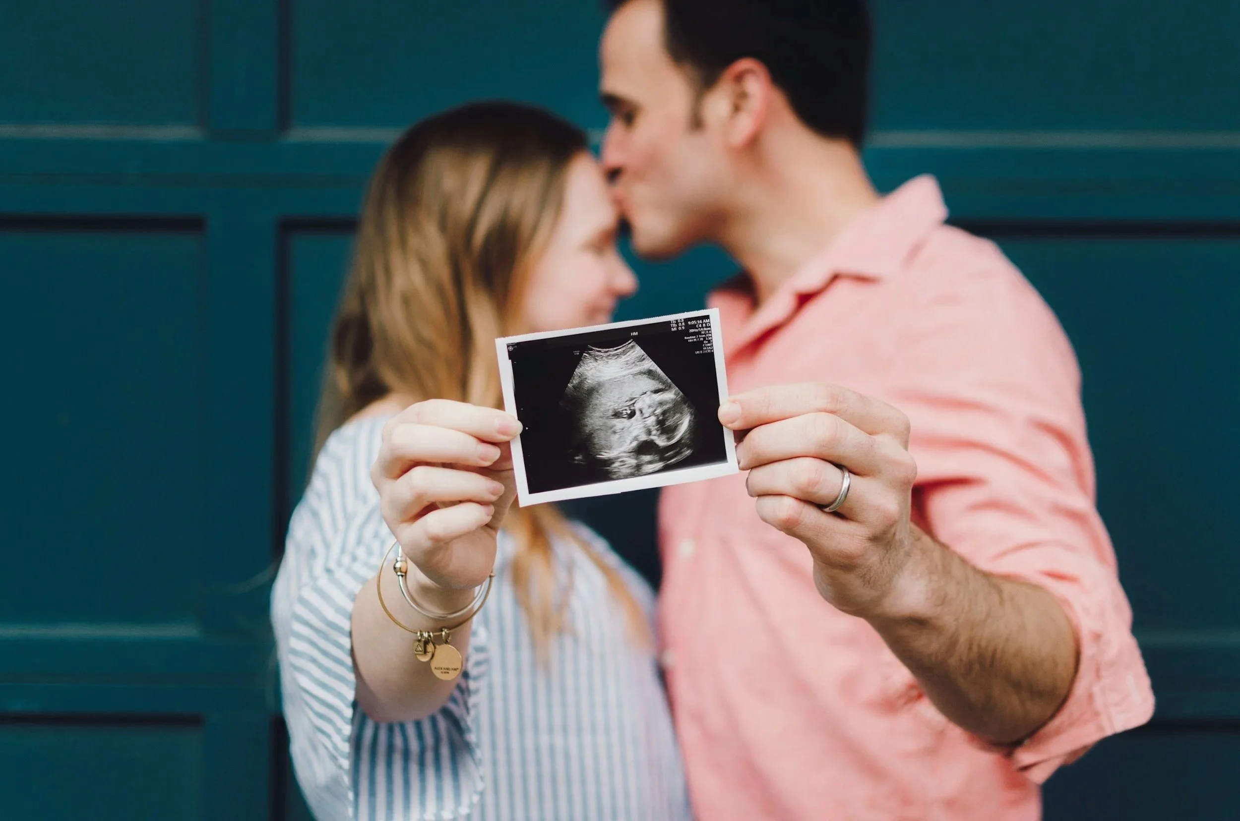 A couple holding an ultrasound image of a baby, with the man kissing the woman on the forehead in front of a blue background.