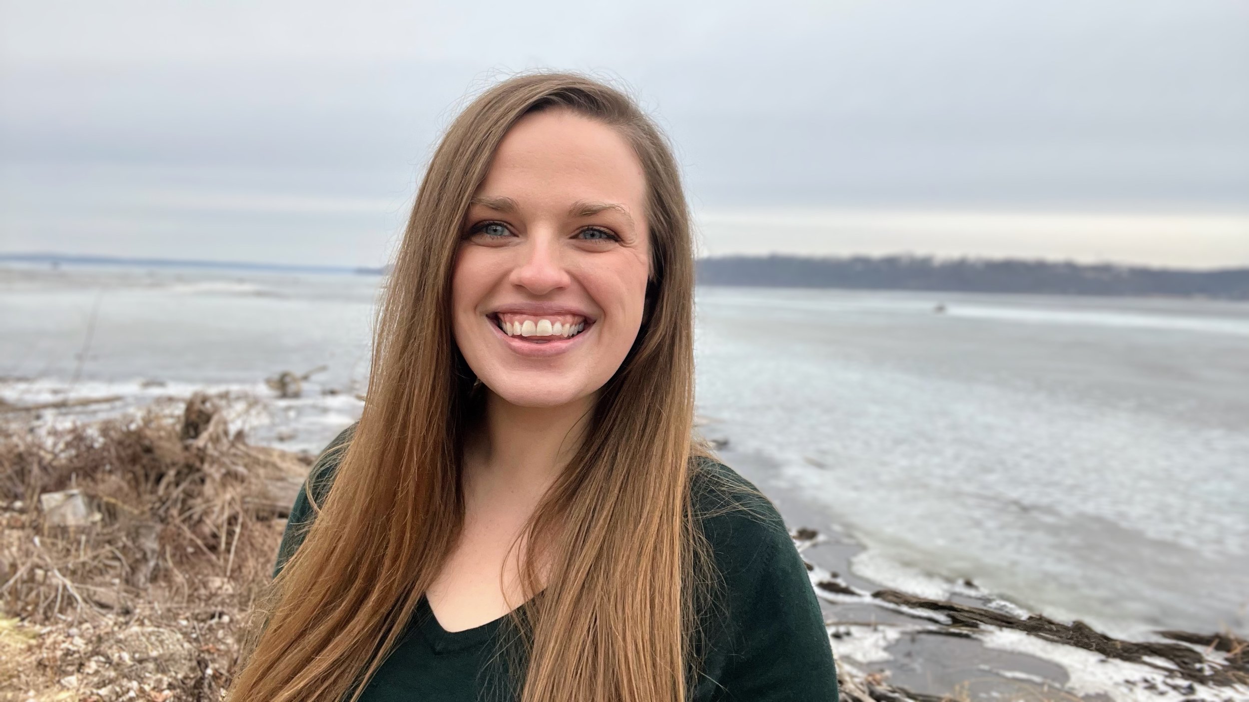 Birth and Postpartum Doula, Stephanie Cook, with long hair standing near a snowy shoreline on a cloudy day.