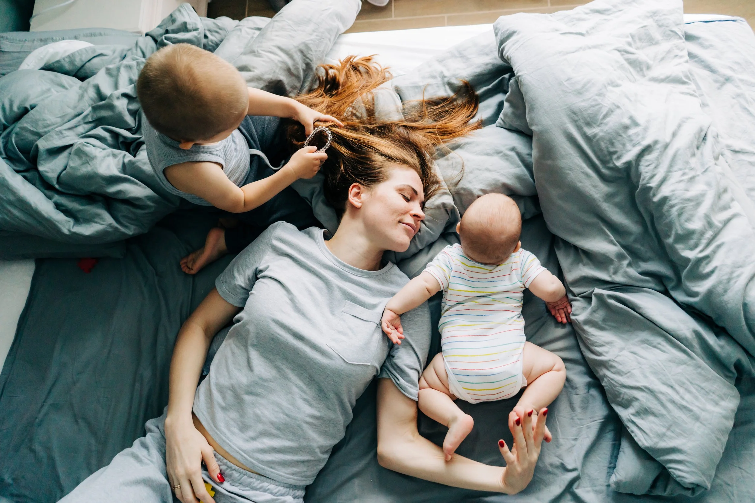A postpartum mother with red hair lying on a bed with two young children, one holding a bracelet, all lying on rumpled gray sheets, appearing relaxed and happy.