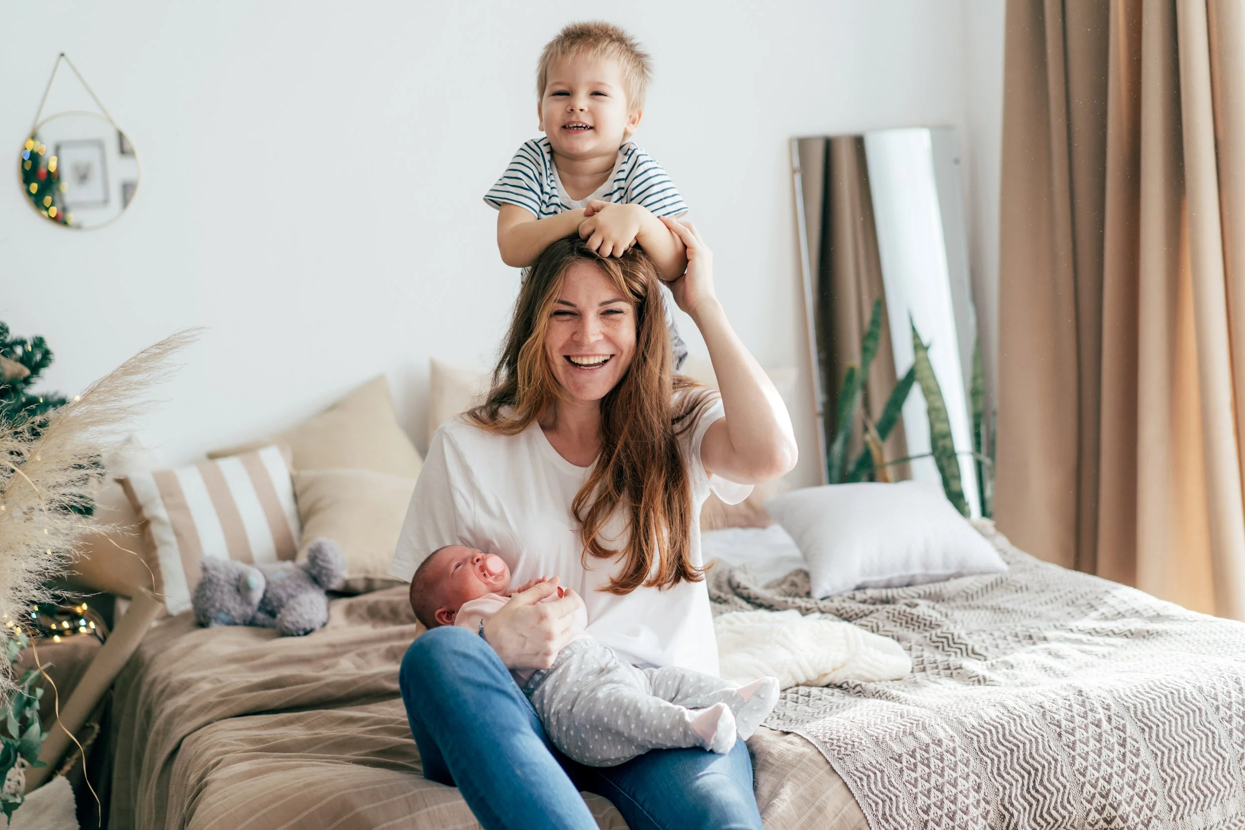 A mother with long brown hair smiling and holding a newborn baby on her lap. A young boy, her son, is standing on her shoulders with arms around her head, smiling. They are in a bedroom with a bed, pillows, and a mirror in the background.
