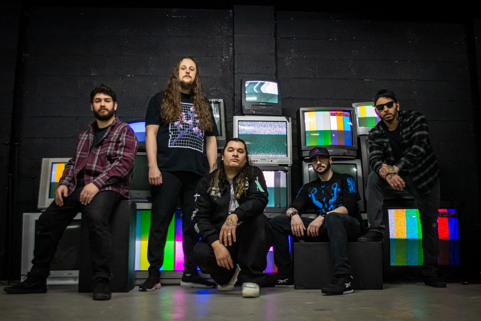 Five men in casual clothing pose in front of a wall of vintage television screens displaying static and color bars, in a dark room.