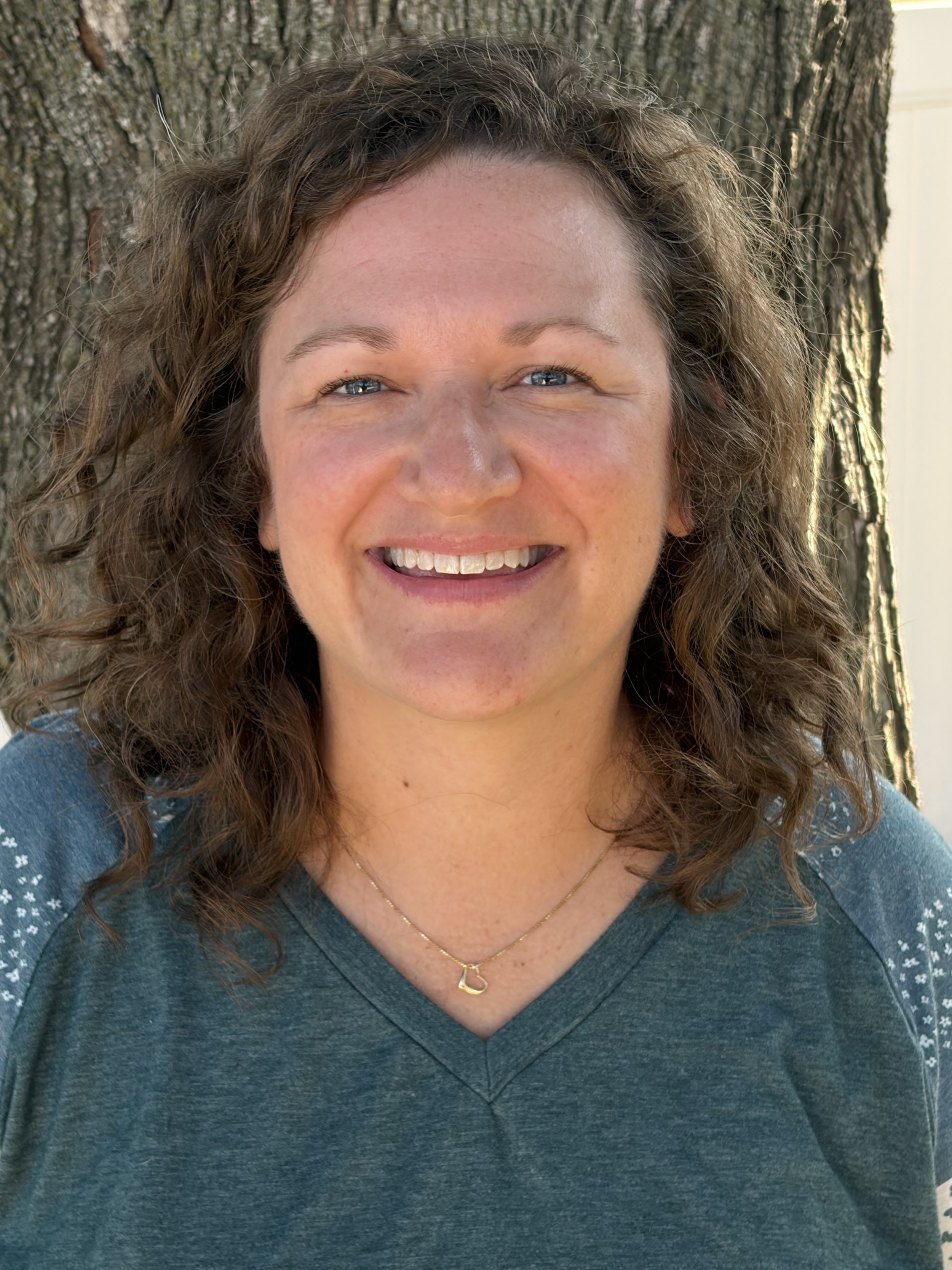 Maura: Brown curly hair, blue-green shirt, blue eyes. Sitting near a tree and smiling. 