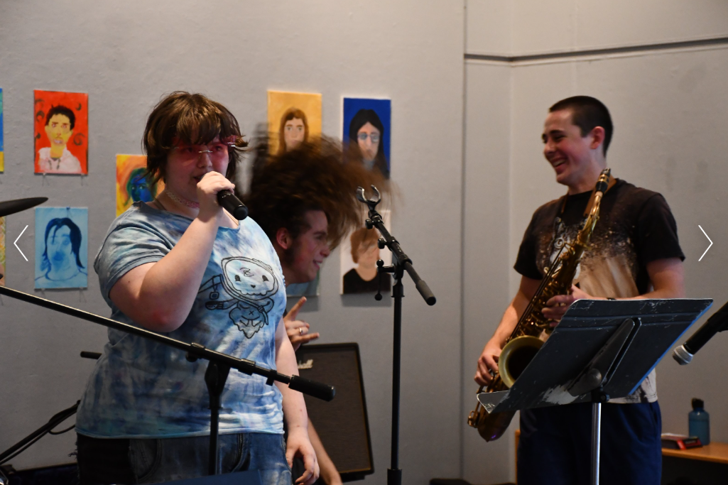 A group of young musicians performing in a room with colorful portrait paintings on the wall. One person with glasses and a Tie-Dye shirt sings into a microphone, while a boy with a saxophone smiles nearby.