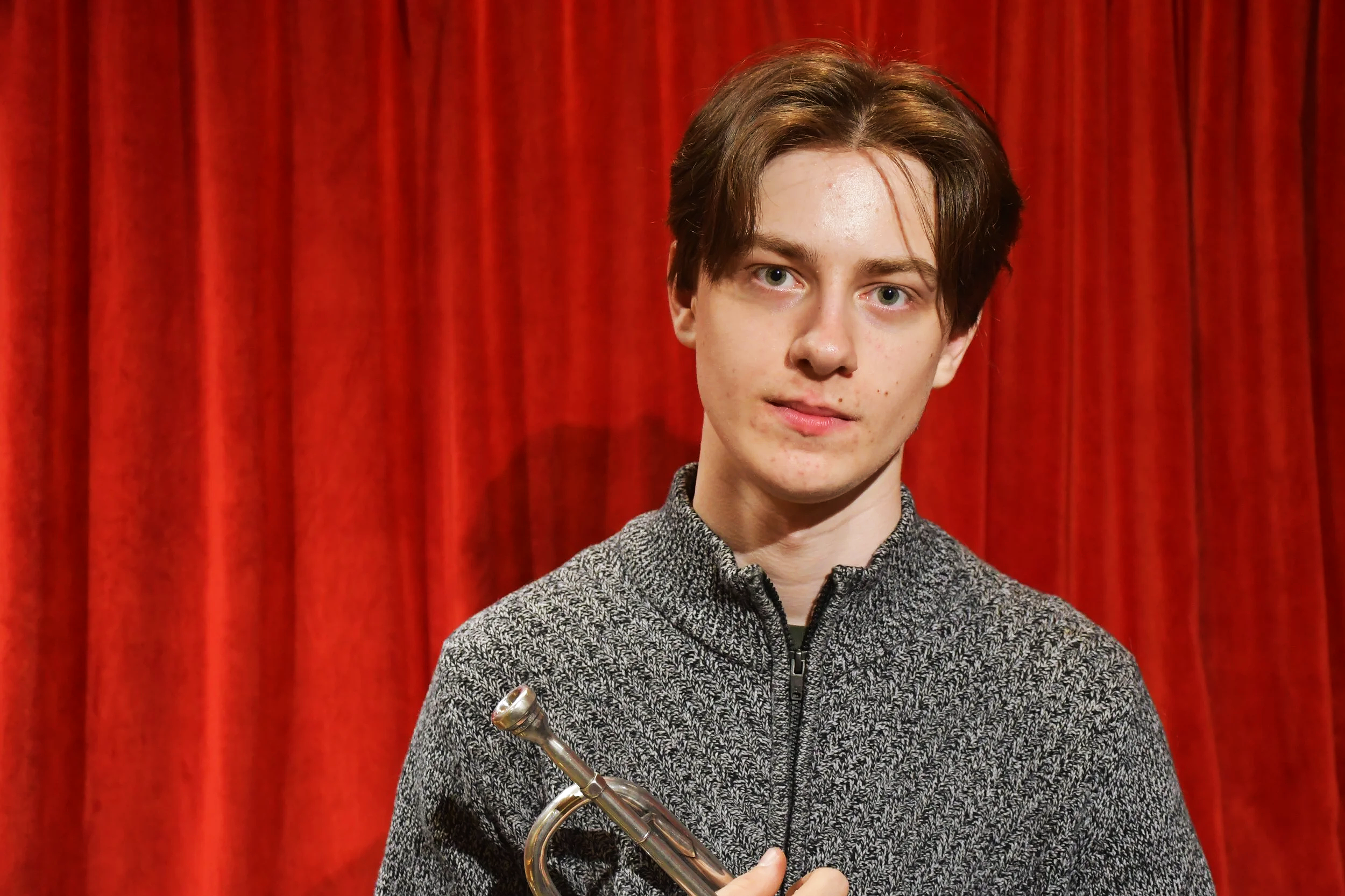 A young man with brown hair holding a trumpet in front of a red curtain.