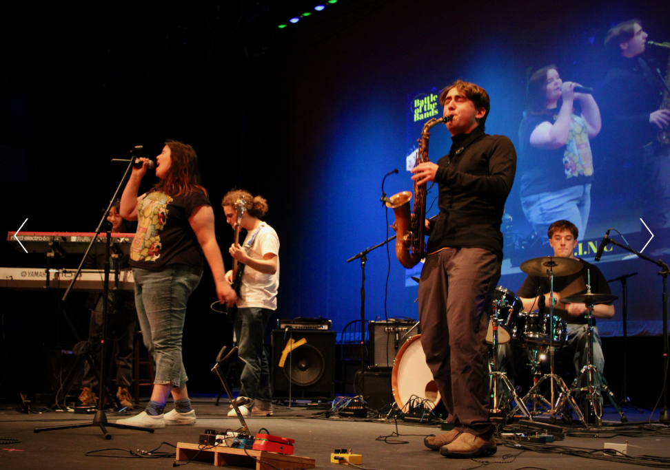 A band performs on stage with a keyboard, guitar, saxophone, drums, and vocals. A large screen behind them displays their image.
