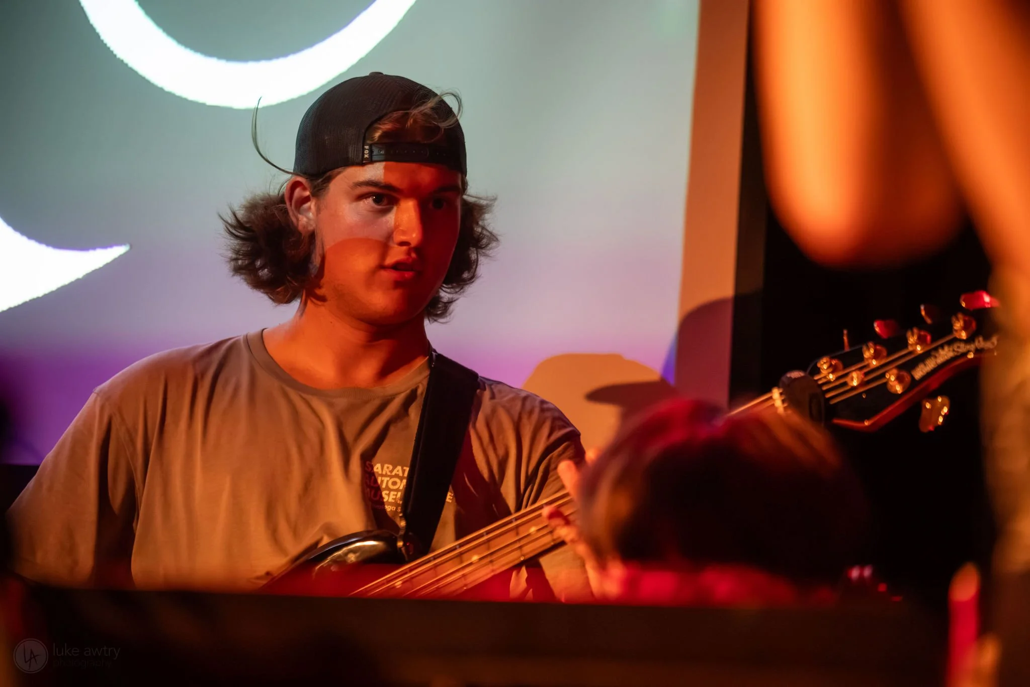 Young man with long hair wearing a baseball cap and black hoodie, holding a bass guitar, standing in front of red curtains.