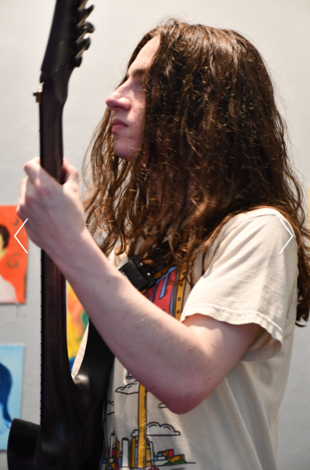 A woman with long, curly brown hair playing an electric guitar in a room decorated with colorful artwork.