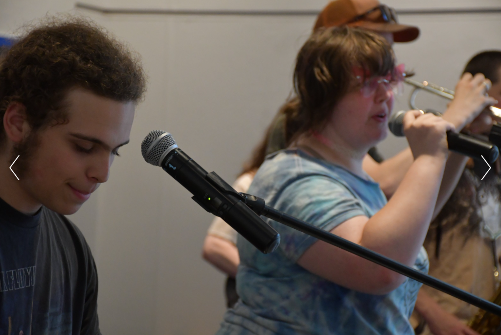 A group of young people performing music with microphones, including a person playing the trumpet and a girl singing or speaking into a microphone.
