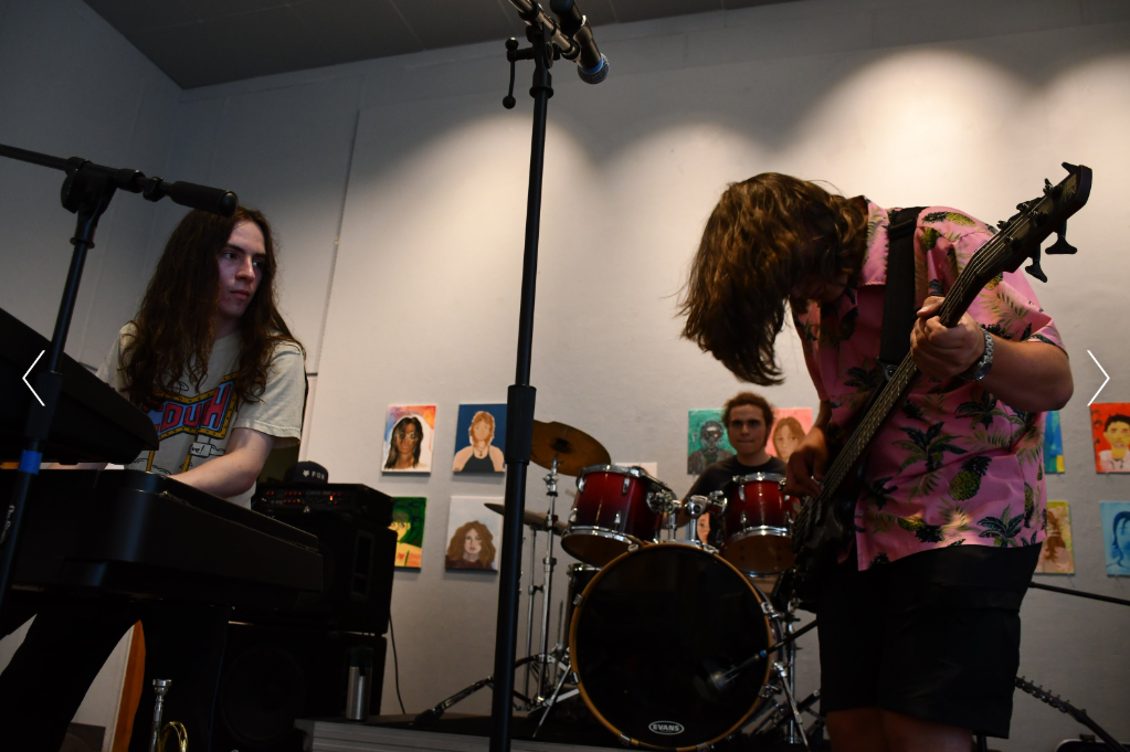 Three young musicians performing in a room with colorful artwork on the wall, including a keyboard player, a drummer, and a guitarist.
