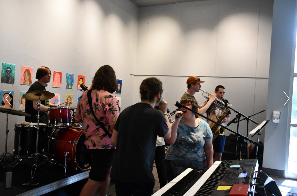 A group of musicians performing in an art gallery with colorful portraits on the wall. The band includes a drummer, a keyboardist, and trumpet and saxophone players.