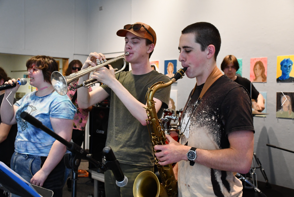 Young people performing music in a room with colorful artwork on the walls, including a person singing into a microphone, two boys playing trumpet and saxophone.