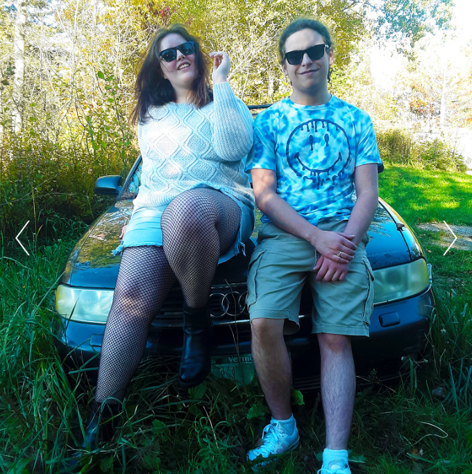 A woman and a man sitting on the hood of a black car outdoors, with green grass and trees in the background, both wearing sunglasses and casual clothing, enjoying a sunny day.