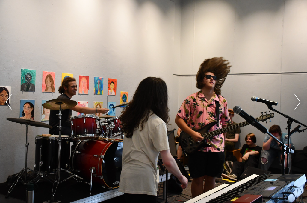 A band performing on a stage with a drummer, guitarist, and keyboardist in front of an audience, with colorful portrait paintings on the wall in the background.