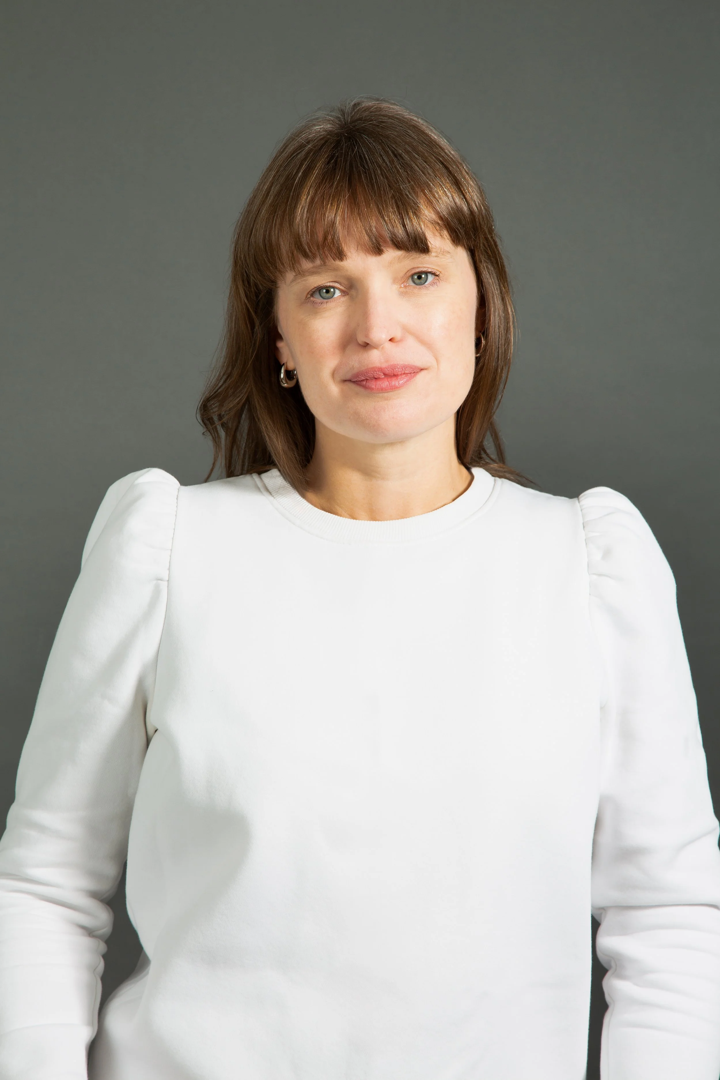 A woman with brown hair, blue eyes, and light makeup wearing a white top with puffed sleeves standing against a gray background.