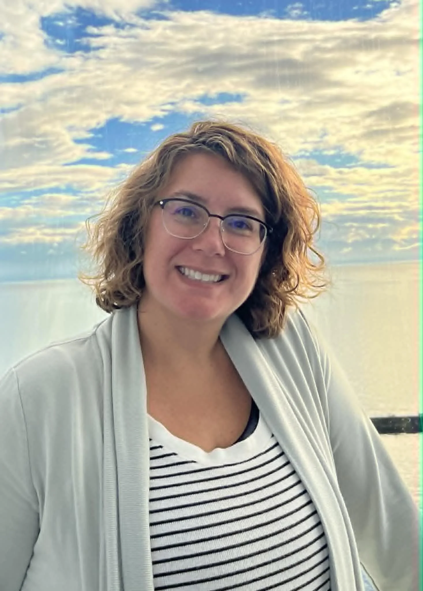 A woman with curly hair and glasses smiling, standing outdoors near a body of water with a sky filled with clouds in the background.