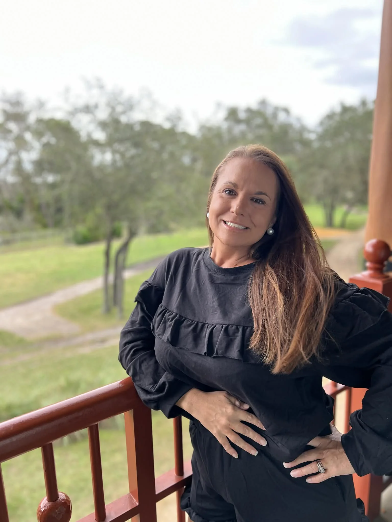 A woman with long brown hair wearing a black top and pearl earrings, standing on a balcony with a red railing, smiling at the camera with a background of trees and a dirt path under a cloudy sky.