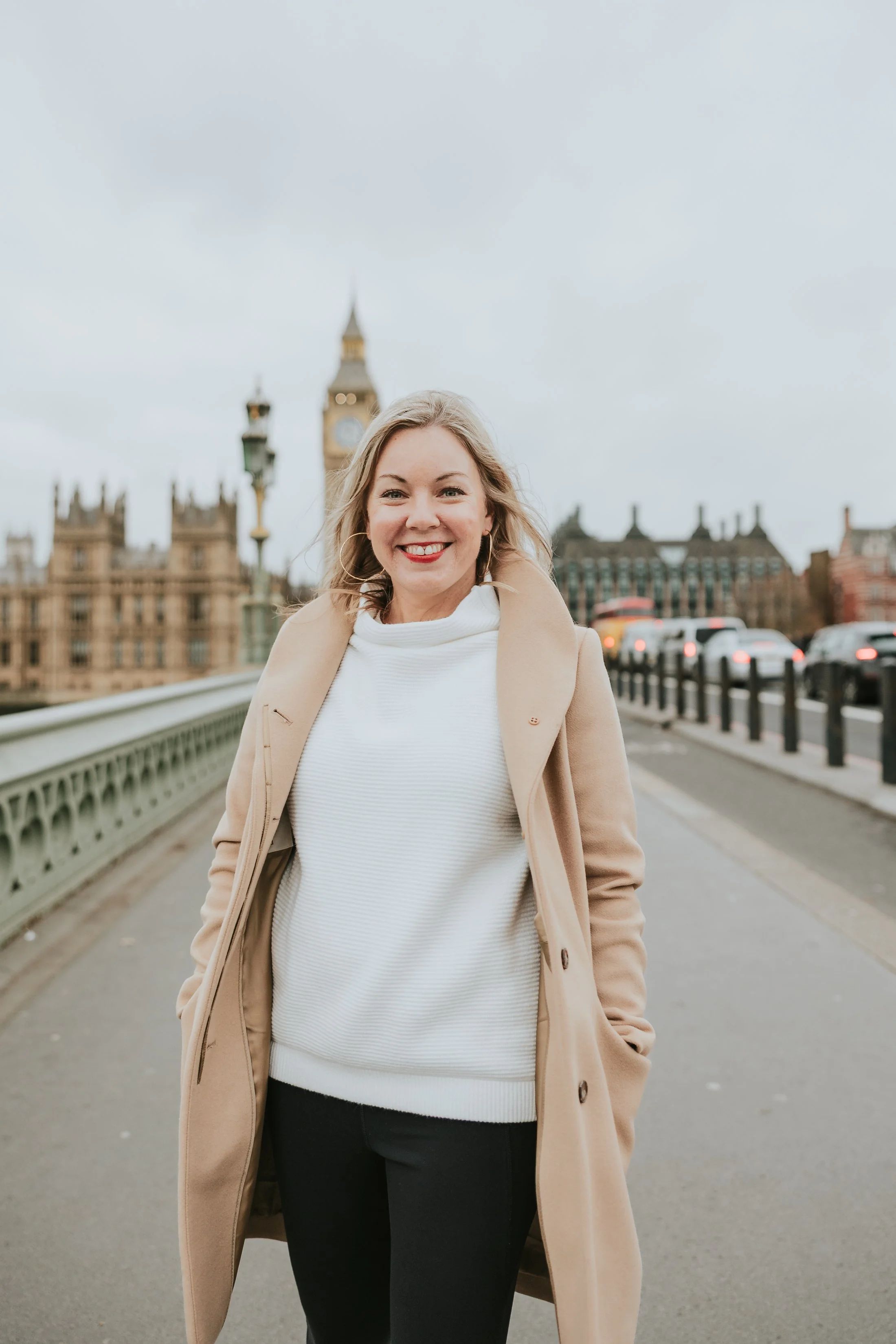 A woman smiling and walking on a bridge, with Big Ben and historic buildings in London in the background.
