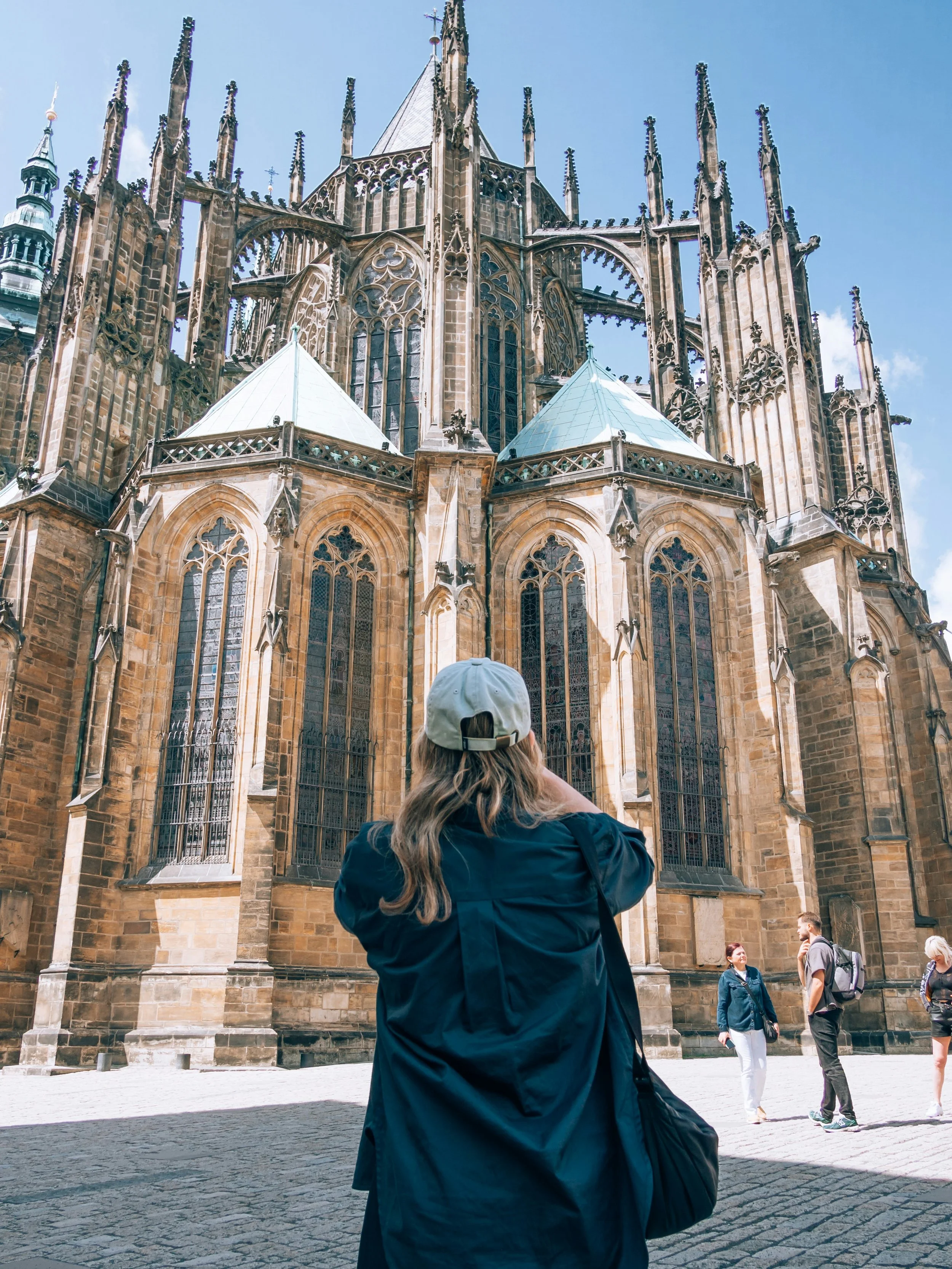 A woman in a blue jacket and a light-colored cap taking a photo of a large, intricate Gothic cathedral with a person looking up at the structure and other visitors nearby.