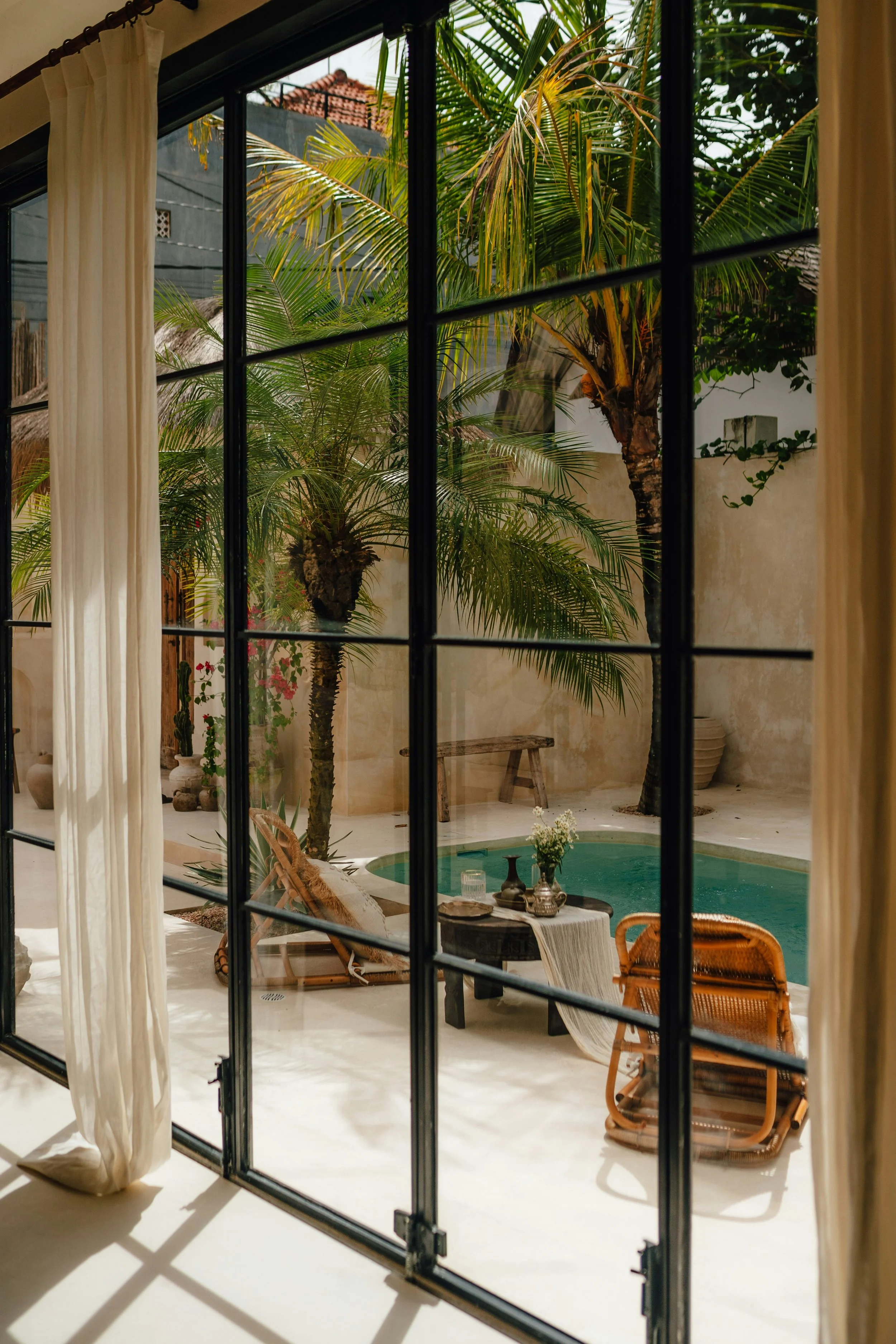View of outdoor patio with small swimming pool, lounge chairs, small table with decorative vases, lush green palm trees, and a bench, seen through a sliding glass door with cream curtains.