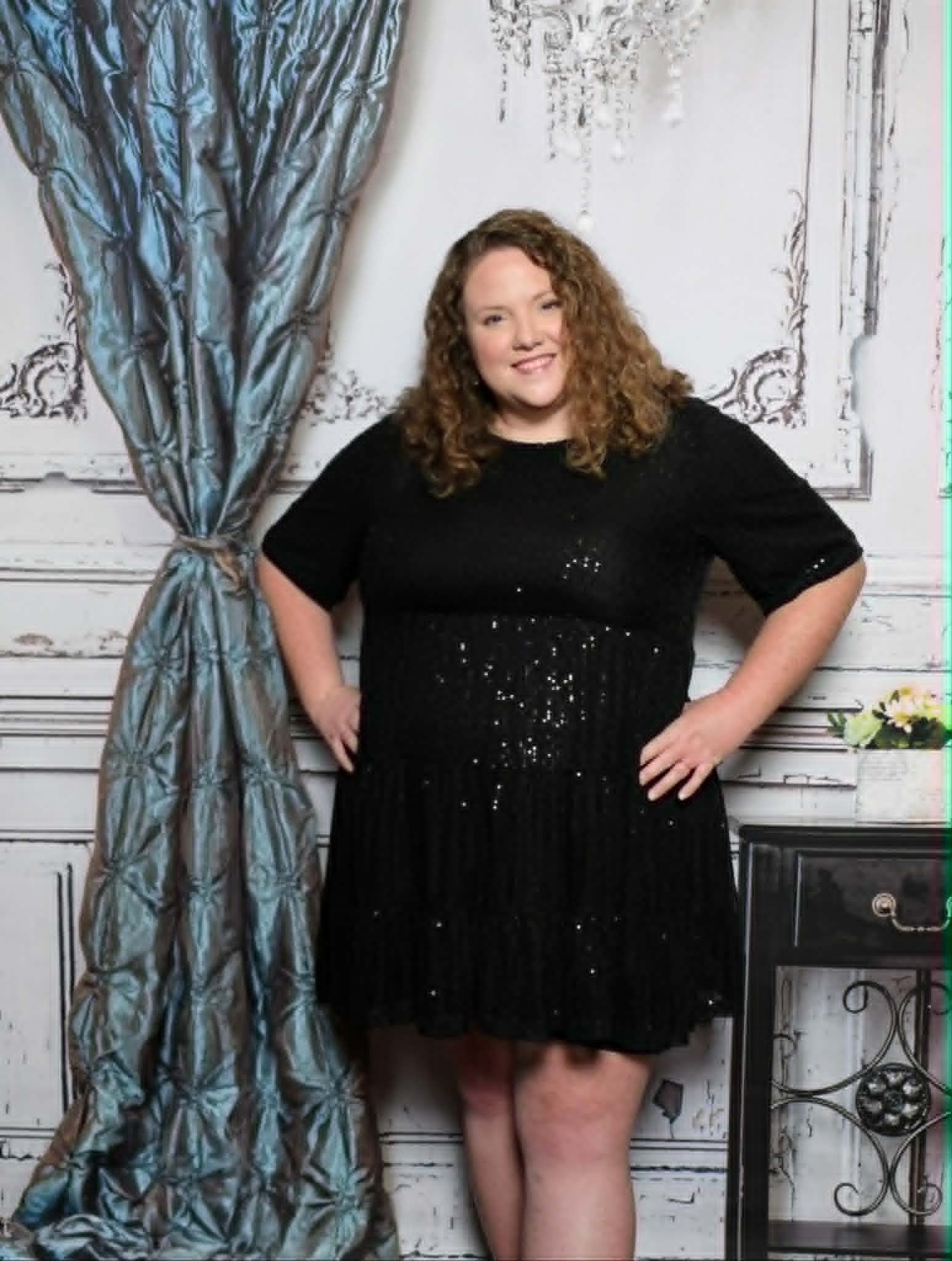 Smiling woman with curly hair wearing a colorful patterned blouse, standing in front of a gold tinsel backdrop with string lights and a black balloon overhead.