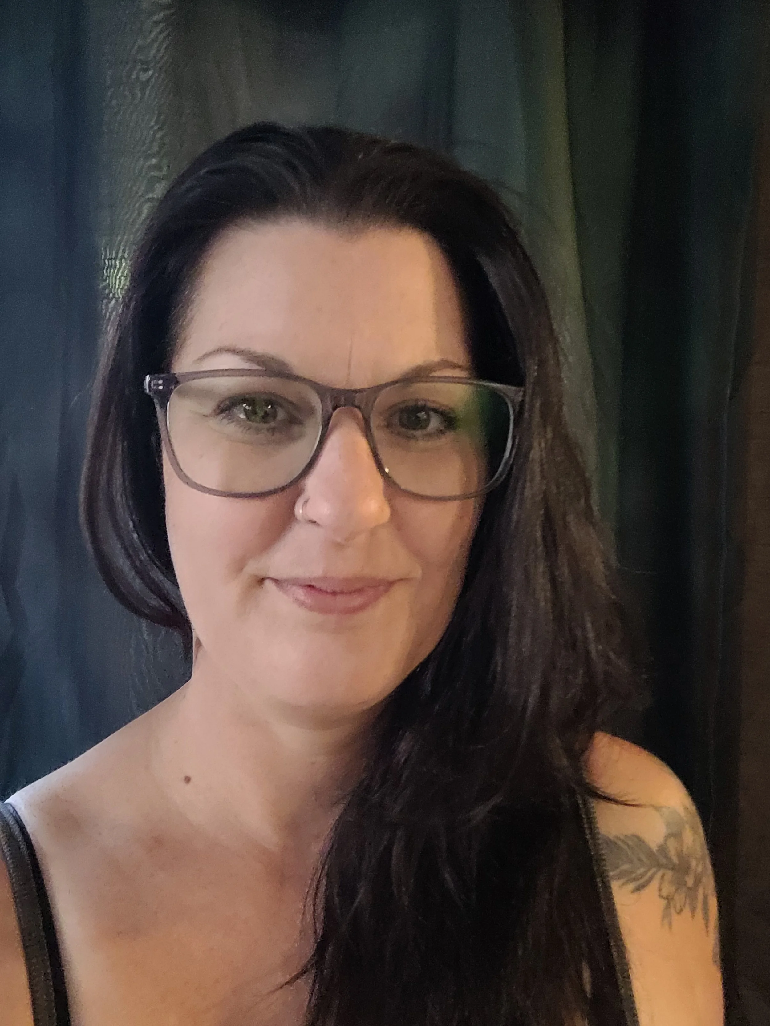 Portrait d'une femme avec des lunettes, cheveux longs noirs, tatouage sur l'épaule, fond sombre.