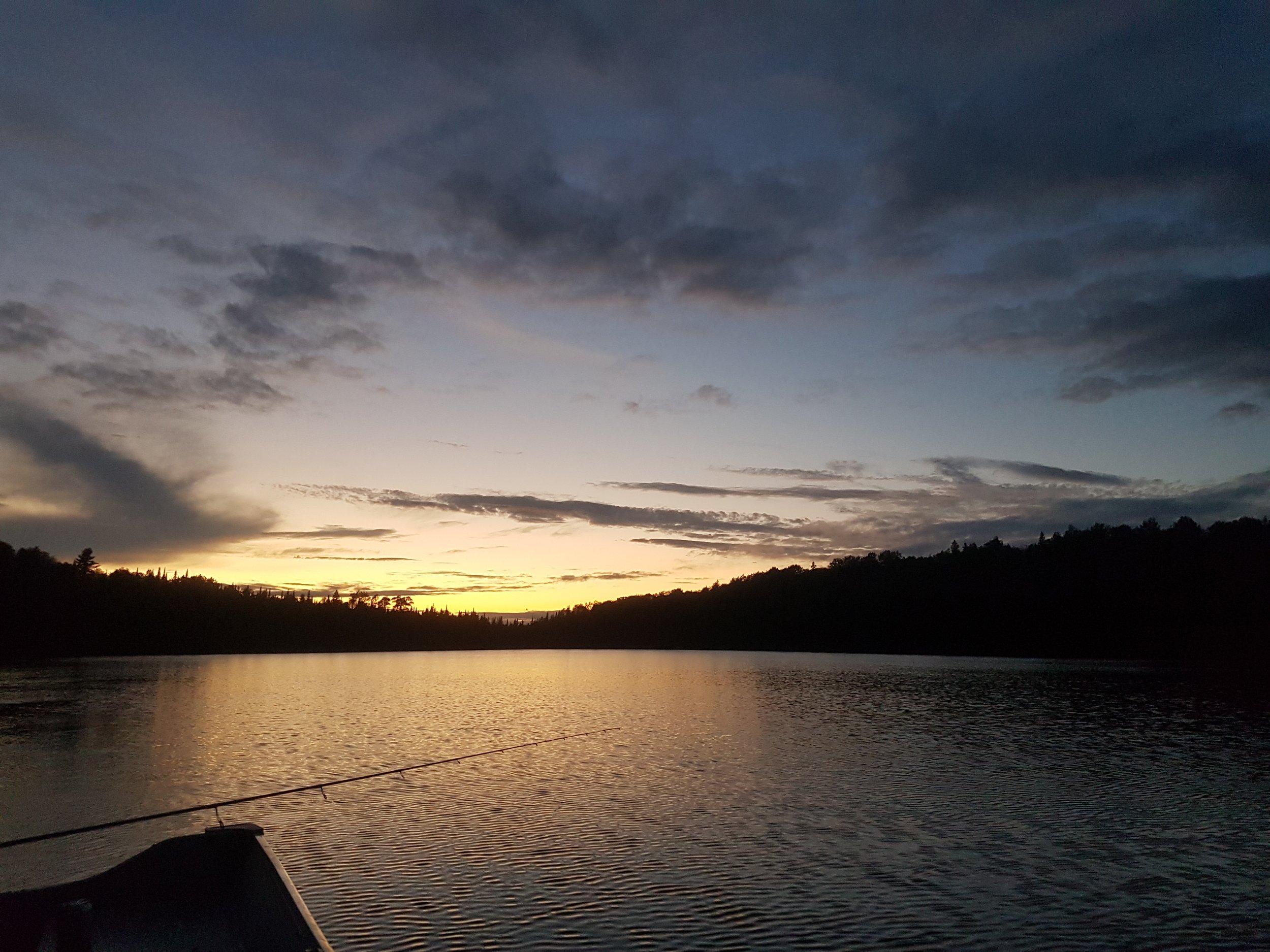 Coucher de soleil sur un lac, avec un ciel partiellement nuageux et une silhouette d'arbres à l'horizon.