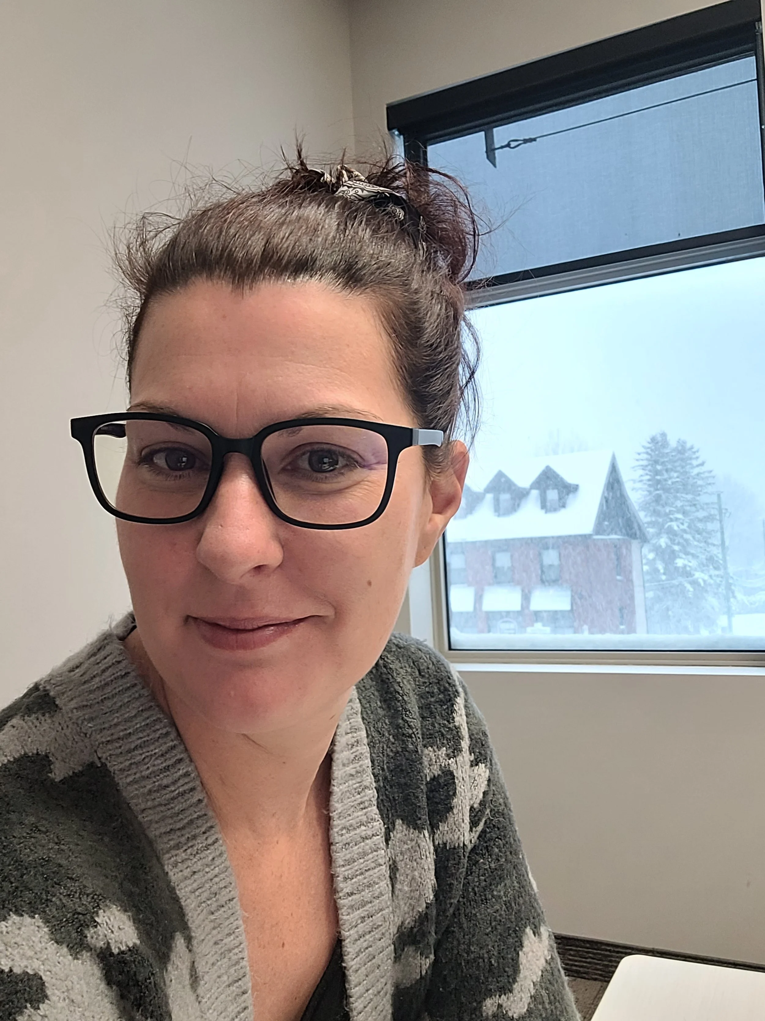 Une femme portant des lunettes noires, souriant légèrement, à l'intérieur d'une pièce près d'une fenêtre avec vue sur une aventure enneigée et des bâtiments.