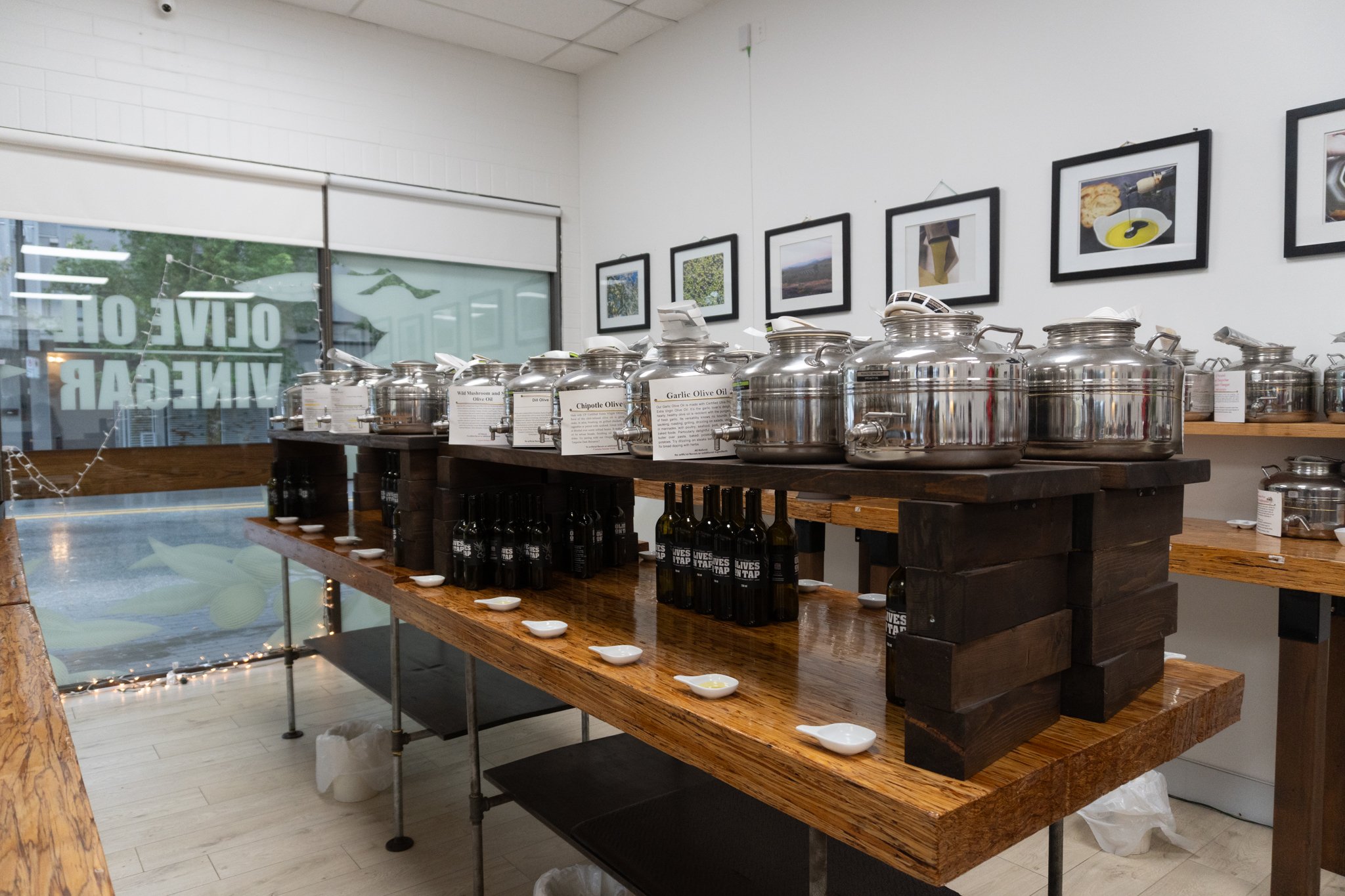 Display of various flavored olive oils in large stainless steel containers on a rustic wooden table in a store. Small tasting spoons with olive oils are arranged in front, and bottles are underneath. Framed pictures hang on the wall behind.