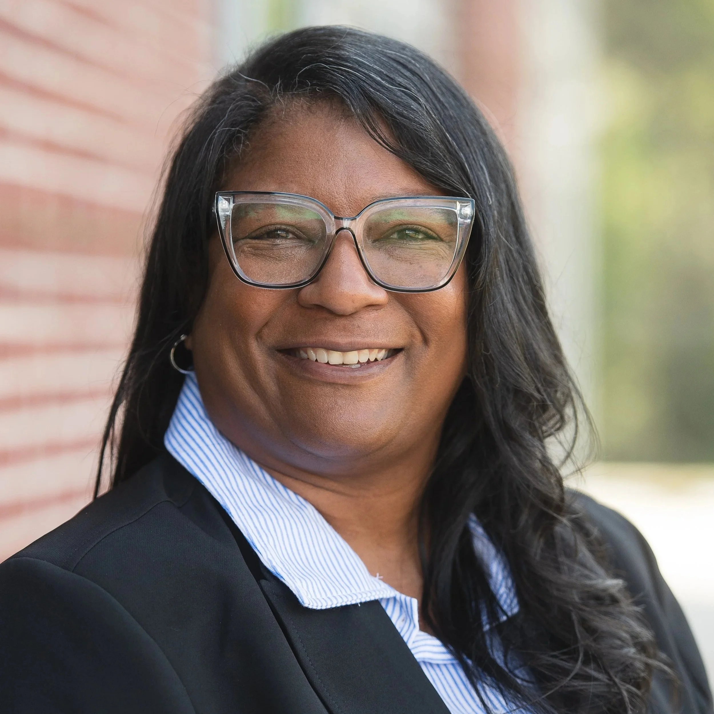 A smiling woman with long black hair, glasses, and a pearl necklace standing in front of a brick wall.