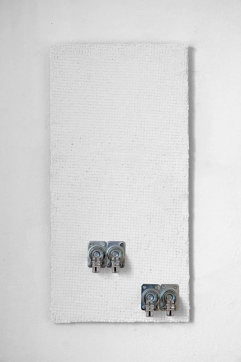 White wall with a white, textured rectangular board mounted on it and four electrical outlets, two on the upper left and two on the lower right.